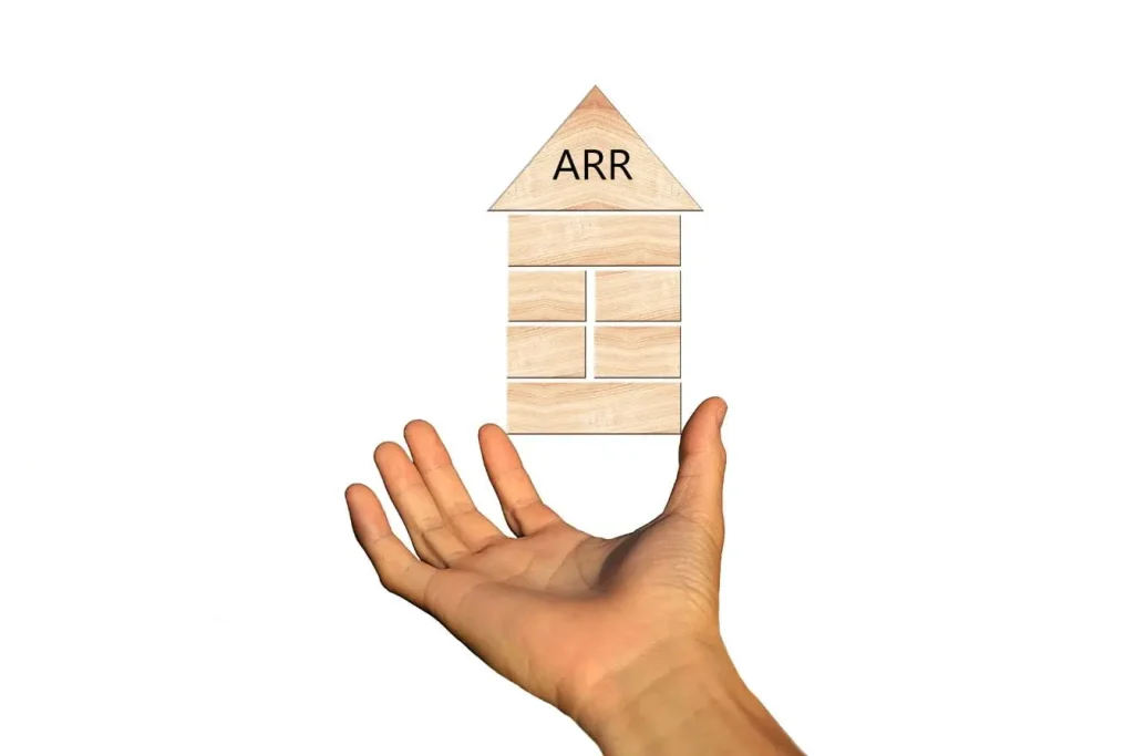 Main tendue vers maison en bois avec ARR inscrit, image illustrant distinction entre revenus mensuels MRR et revenus annuels ARR dans modèle SaaS.