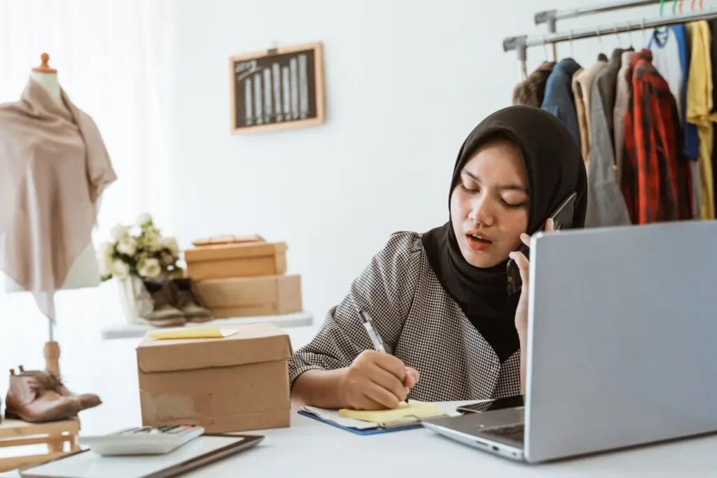 Femme en hijab travaillant sur ordinateur et téléphone, préparant commandes de vêtements, illustrant la vente en dropshipping et la gestion e‑commerce