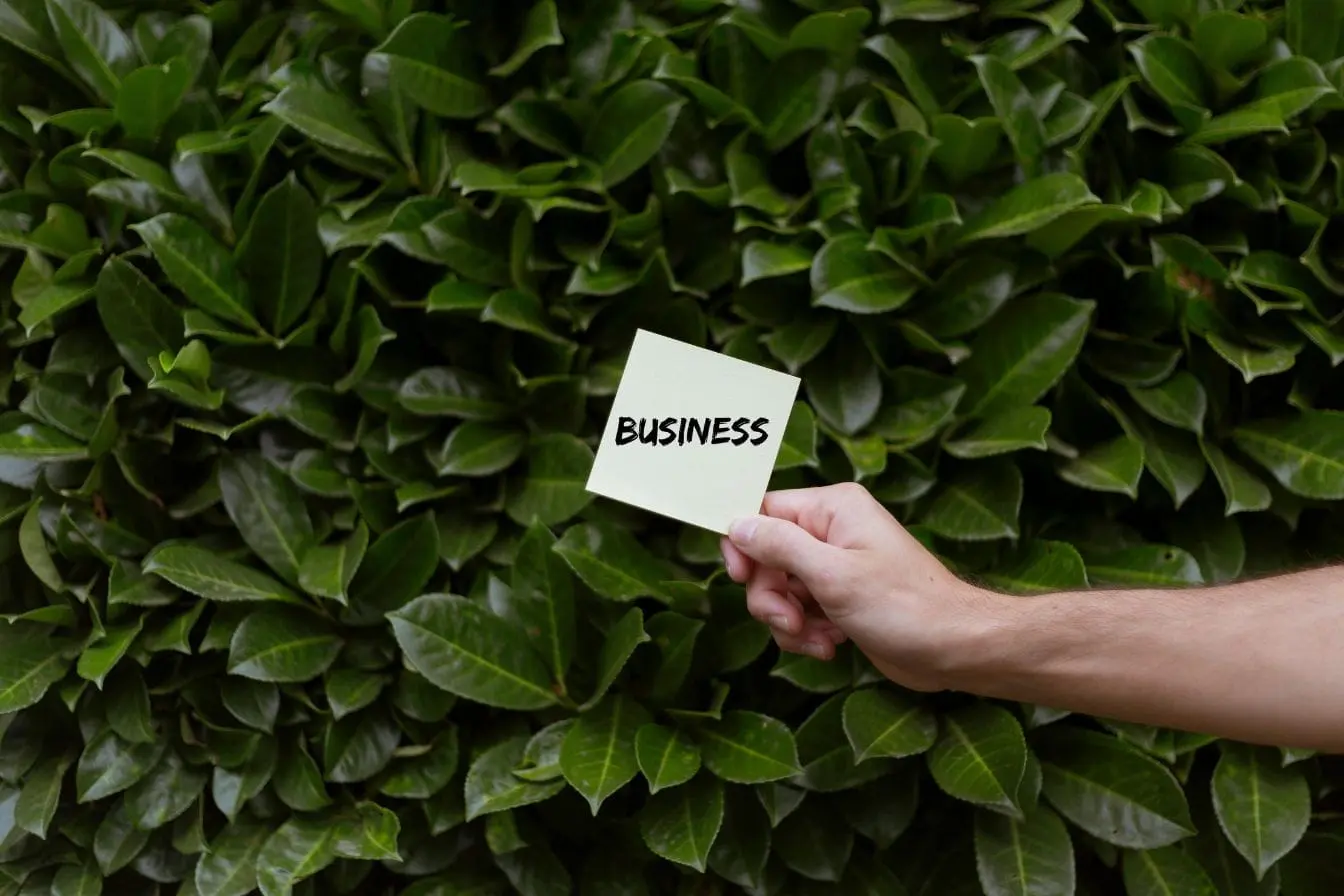 Main tenant un post-it marqué business devant feuillage vert, image illustrant le Green IT et la synergie entre technologie et durabilité.