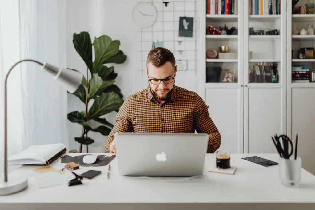 Personne assise à un bureau blanc travaillant sur un MacBook, entourée de carnet, café et outils, image d’un espace créatif simplifié pour non-designers.