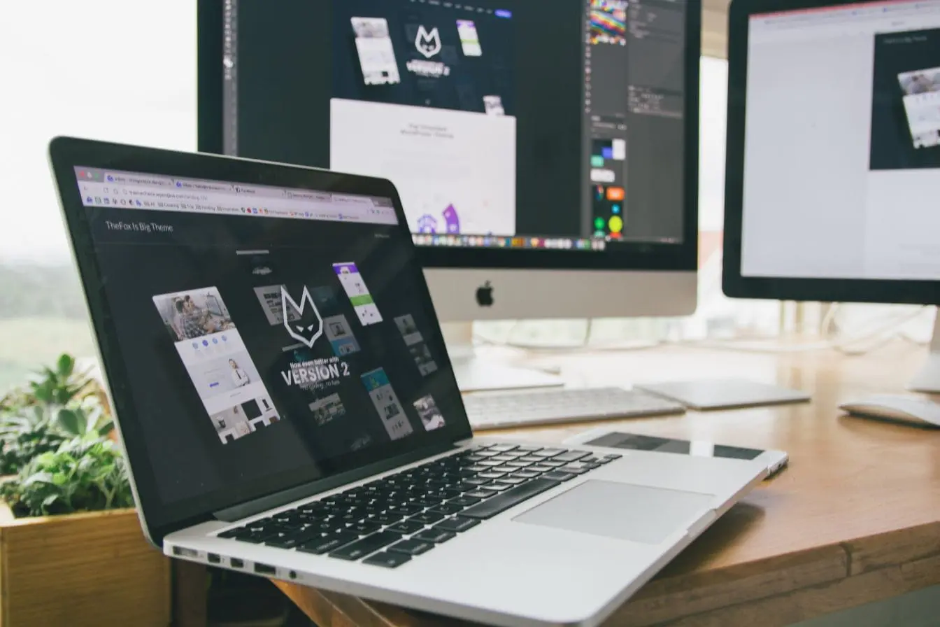 Un bureau moderne avec trois écrans affichant des maquettes web et outils graphiques, illustrant un espace de création simplifié pour non-designers.