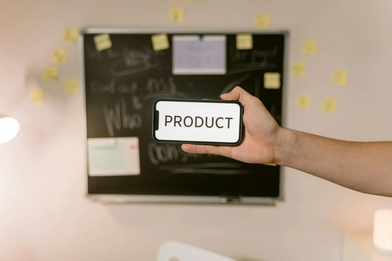 Main tenant smartphone affichant mot PRODUCT, image illustrant la recherche du Product Market Fit dans un environnement de travail créatif.