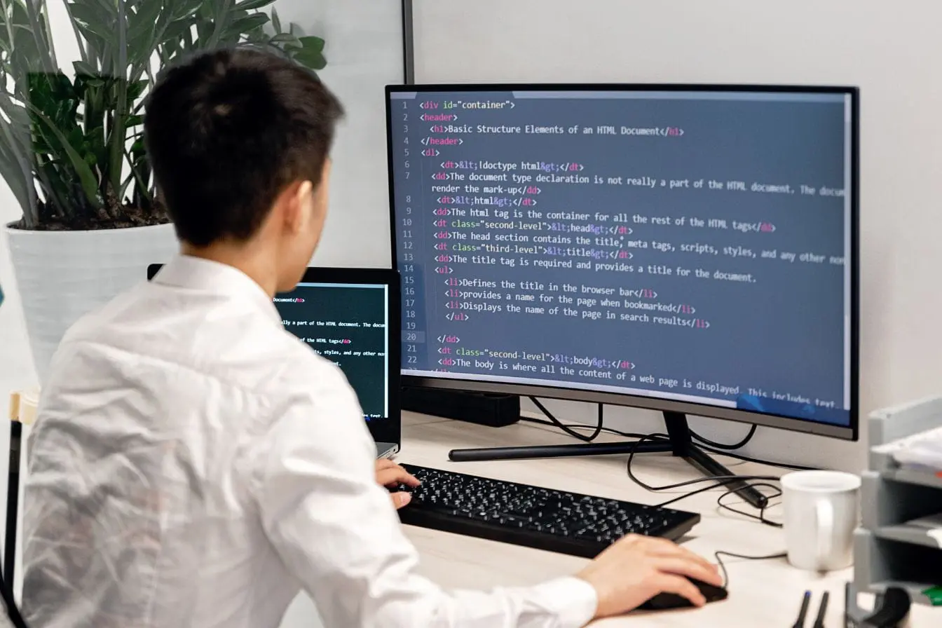 Personne assise devant double écran affichant code HTML, image qui illustre l’univers technique où Kubernetes orchestre les applications.