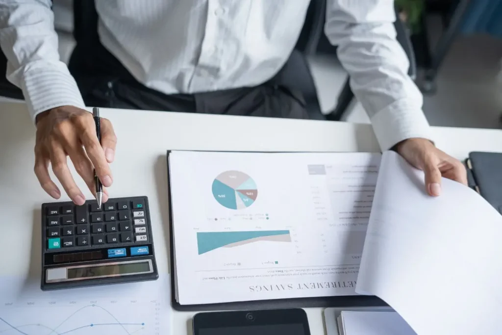 Personne en chemise blanche utilisant calculatrice sur bureau avec graphiques financiers, reflet du calcul du ratio CAC LTV pour mesurer rentabilité client.