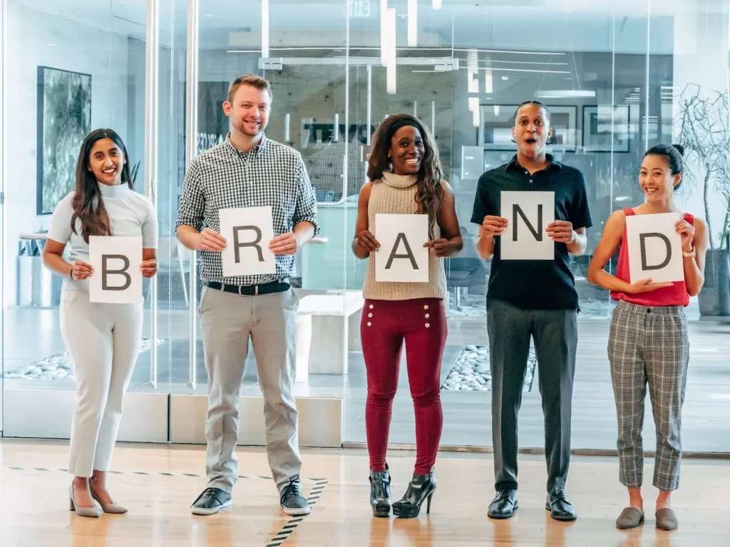 Cinq personnes debout en bureau moderne tenant feuilles formant mot BRAND, symbole de la collaboration et du co‑branding en marketing.