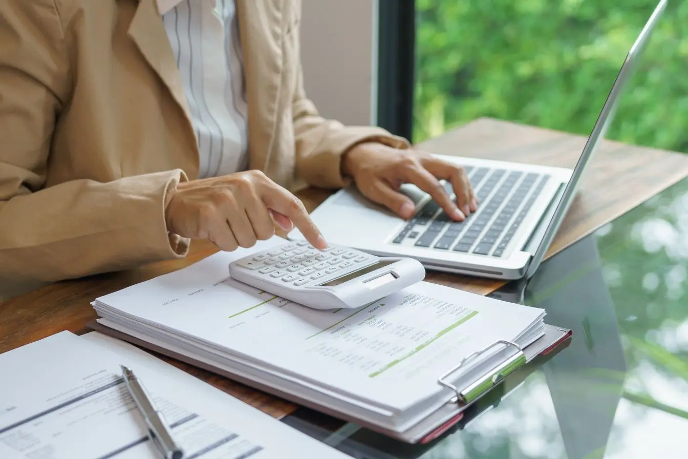 calculer le seuil de rentabilité d’une entreprise Personne en veste beige utilisant calculatrice sur bureau, symbole des pratiques pour calculer le seuil de rentabilité d’une entreprise.