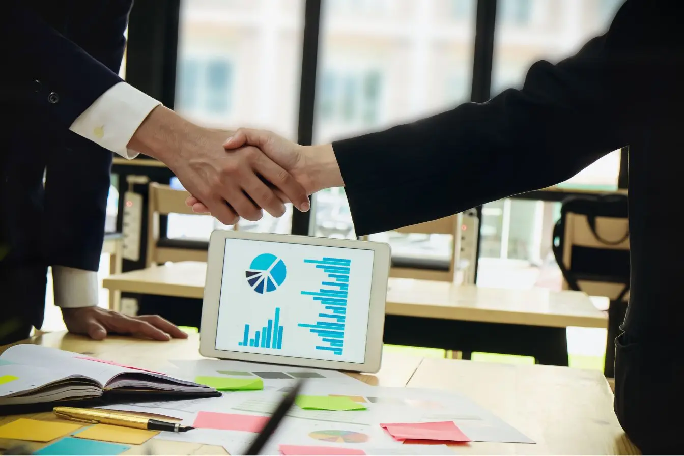joint-venture Deux personnes en costume se serrant la main devant tablette affichant graphiques, symbole d’un accord professionnel en joint-venture.