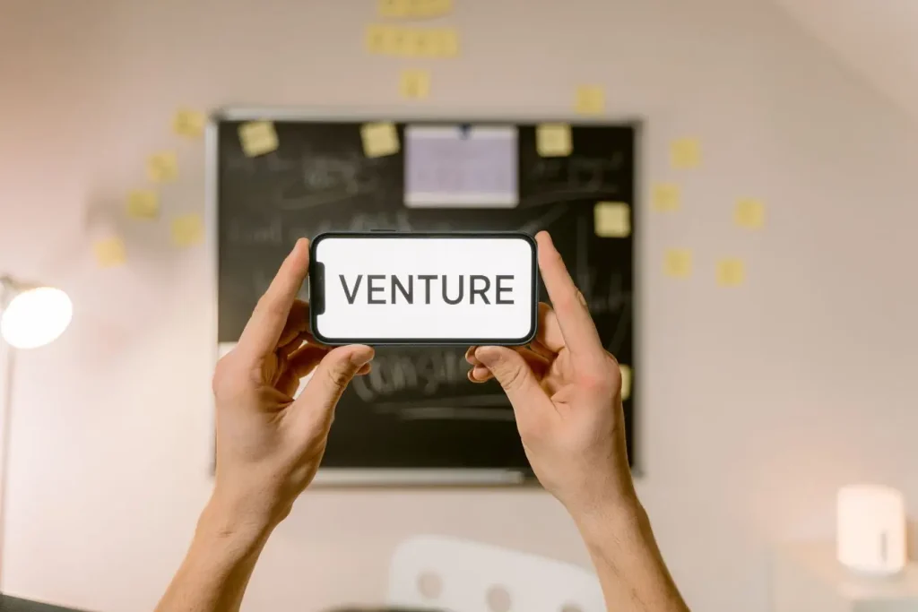 Deux mains tenant smartphone affichant mot Venture, symbole d’un accord professionnel en joint-venture.